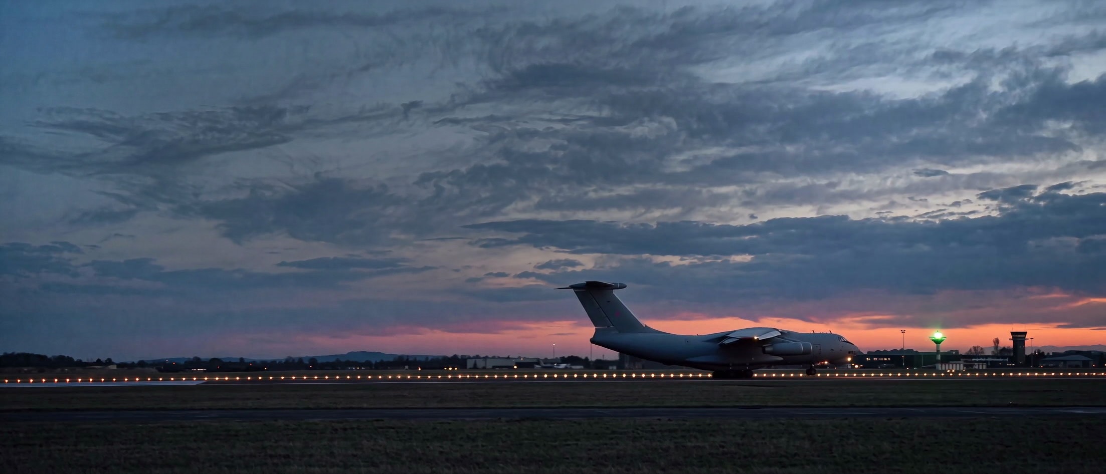 A grey military transport on the runway at blue hour just after landing, rolling out past the control tower under dramatic dusk clouds with the runway edge lights glowing
